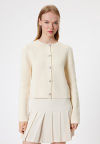 Crème textuur cardigan met lange mouwen, ronde halslijn en zes decoratieve knopen. Gepaard met een beige geplisseerde rok.