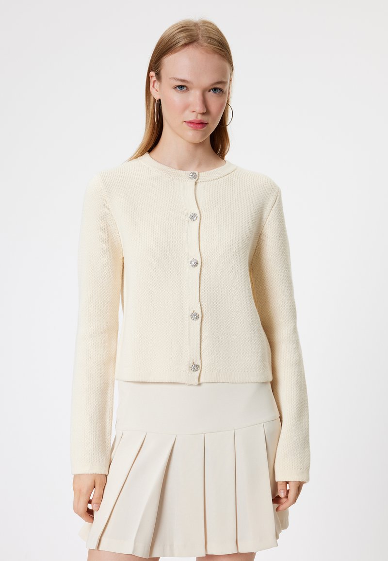Crème textuur cardigan met lange mouwen, ronde halslijn en zes decoratieve knopen. Gepaard met een beige geplisseerde rok.