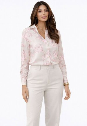Femme aux longs cheveux bruns portant un chemisier rose pâle à fleurs à boutons et un pantalon beige, accessoirisée d'une montre en or et d'un collier chaîne.