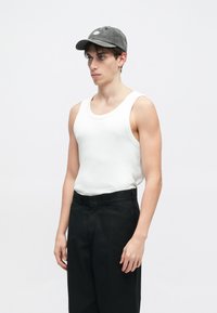 Ung mann iført en hvit tanktop, svarte bukser, grå caps og liten hoop-ørering, som står mot en enkel lys bakgrunn.