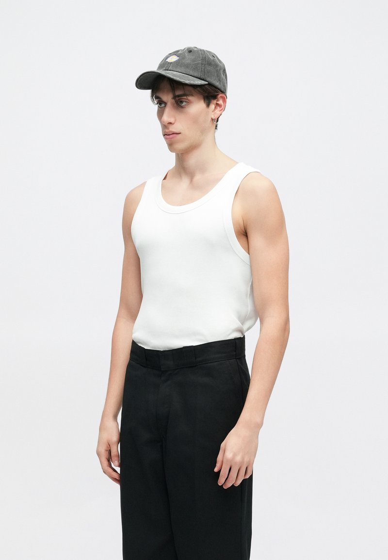 Junger Mann, der ein weißes Tanktop, schwarze Hose, graue Mütze und kleinen Ohrstecker trägt, steht vor einem einfarbigen hellen Hintergrund.