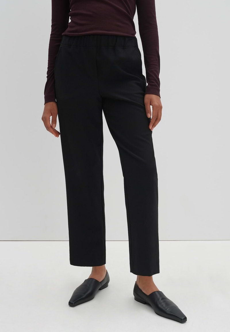 Pantalon noir en design droit, fabriqué en tissu lisse avec une taille élastique, porté avec des chaussure à enfiler noires à bouts pointus.
