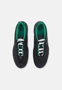 Baskets noires en daim et en mesh avec des lacets blancs et verts, présentant un bout arrondi et une semelle épaisse. Doublure intérieure verte en accent.