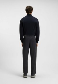 Black knit sweater with a collar and contrasting stripe, paired with dark gray pants featuring pockets. Model stands facing away.