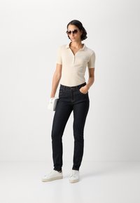 Krämfärgad ribbad polo med korta ärmar, kombinerad med mörkblå skinny jeans, vita sneakers och en vit handväska. Tillbehör inkluderar solglasögon.