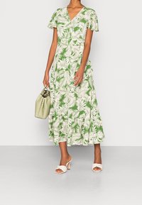 Woman wearing green and white tropical leaf print midi dress, holding a light green handbag, and wearing white braided heeled sandals.