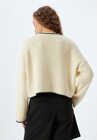 Maglione all'uncinetto di colore crema con un design cropped, rifiniture nere, maniche ampie e orlo a coste. Abbinato a pantaloni neri.