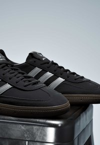 Baskets noires avec des rayures grises et des semelles marron posées sur une surface métallique sur un fond gris uni.