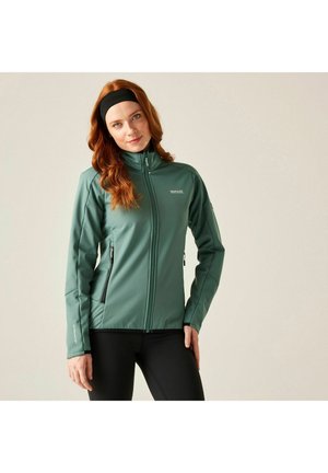 Mujer con cabello rojo que lleva una chaqueta verde con cremallera y leggings negros, posando con una mano en la cadera contra un fondo neutro.