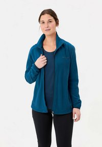 Pruunide juustega naine, kes kannab pealuu värvi lukkuga fliisjacki üle tumesinise särgi ja mustade pükstega, seisab valge tausta ees.