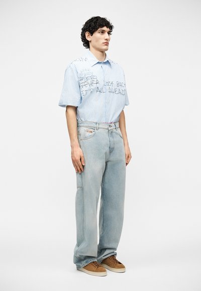 Jeune homme debout, portant une chemise à manches courtes à rayures bleu clair avec du texte, un jean ample délavé clair et des baskets marron sur fond blanc.
