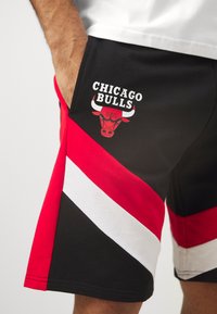 Črne kratke hlače z rdečimi in belimi diagonálnimi črtami, z logotipom Chicago Bulls v beli in rdeči barvi blizu žepa. Narejene iz mehkega materiala.