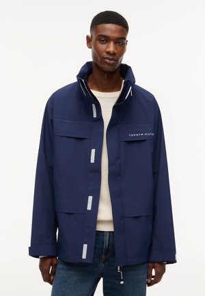 Mand iført marineblå Tommy Hilfiger jakke over hvid sweater og blå jeans, stående mod en ensfarvet hvid baggrund.