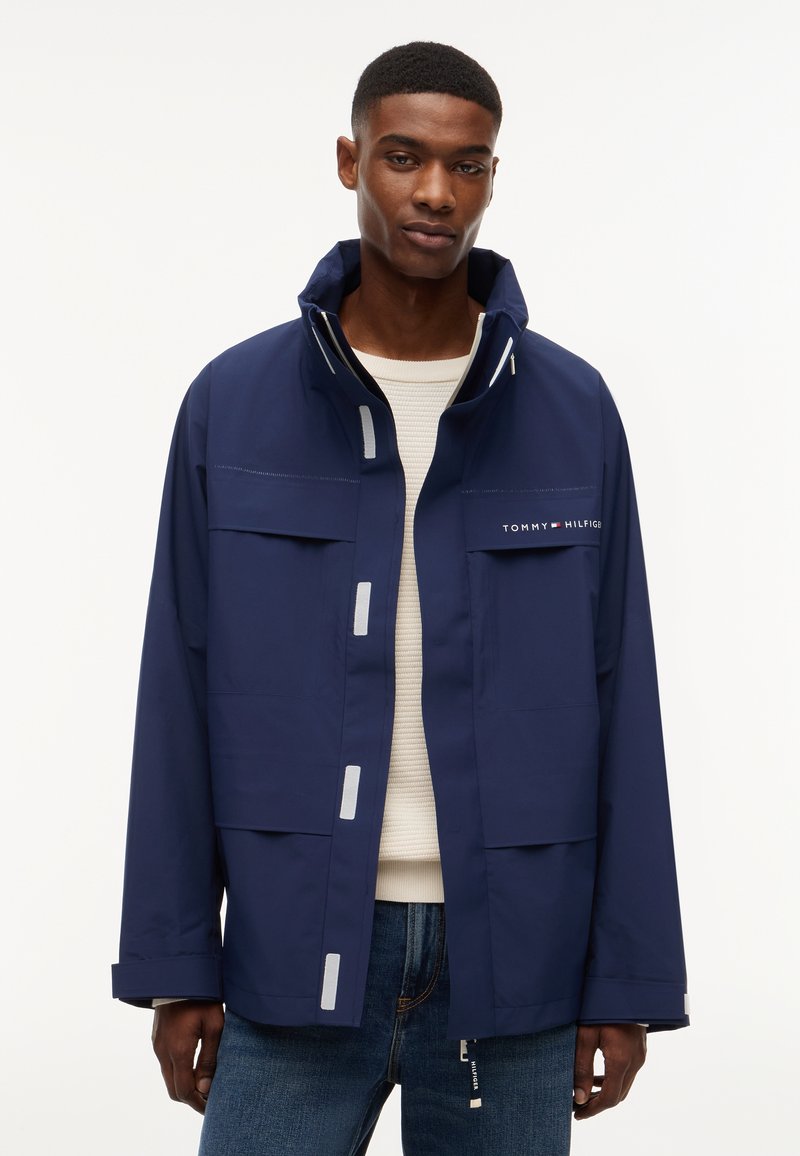 Άνδρας που φοράει μπλε ναυτικό μπουφάν Tommy Hilfiger πάνω από λευκό πουλόβερ και μπλε τζιν, στέκεται μπροστά σε απλό λευκό φόντο.