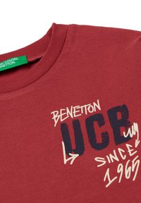 Czerwona koszulka bawełniana z okrągłym dekoltem. Zawiera biały i granatowy tekst: "BENETTON UCB od 1965 roku" z graficznymi akcentami oraz zieloną metką.