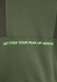 Felpa verde con una texture liscia, caratterizzata da un pannello verde scuro a contrasto e un testo audace in bianco e verde neon: "SUPERARE LA PAURA DELL'ALTEZZA."