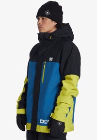 Kleurijke skijas in blauw, zwart en felgeel. Voorzien van een rits aan de voorkant, een capuchon en een logo op de borst, met contrasterende accenten op de mouwen.