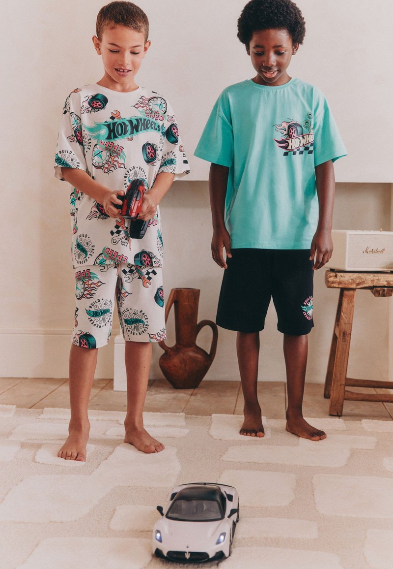 To børn står på et tekstureret tæppe, den ene iført en hvid T-shirt med grafik og shorts, den anden en ensfarvet turkis T-shirt og sorte shorts. En legetøjsbil står foran dem.