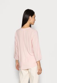 Blusa rosa chiaro con maniche a tre quarti, scollatura rotonda e dettaglio di piega sul retro, realizzata in un tessuto morbido e liscio. Indossata con pantaloni chiari.