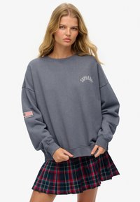 Jonge vrouw met lang blond haar die een grijze Superdry sweatshirt en een geplooide gestreepte rok in rood, marineblauw en groen draagt, tegen een witte achtergrond.