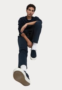 Marineblaue Sweatshirt und Hose, getragen von einer sitzenden Person, mit einem Paar marineblauer Slipper mit weißer Sohle und strukturiertem Gummiuntergrund.