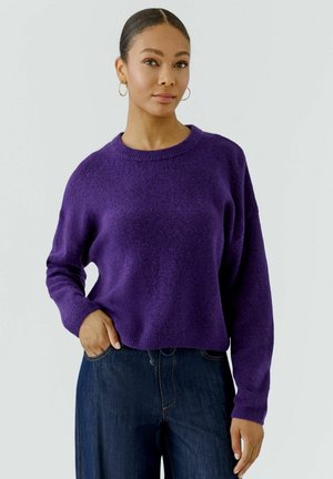 Vrouw met het haar naar achteren gedragen draagt creolen, een paarse trui met lange mouwen en donkerblauwe spijkerbroek, staand met één hand in de zak.