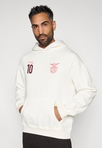 Sudadera de algodón blanca con capucha ajustable, que presenta detalles bordados en rojo: "10" y un escudo. Incluye un bolsillo frontal tipo canguro.