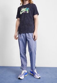 Person wearing a black Nike T-shirt with green palm tree graphic, light blue cargo pants, and white sneakers with blue and neon green accents.