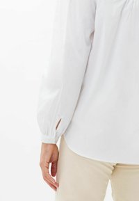 Blusa blanca con mangas abullonadas y puños con botones, con cortes laterales y terminada en un dobladillo redondeado. Textura de tela suave.