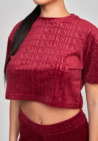 Top corto de terciopelo rojo con patrón de logo en relieve, mangas cortas y escote redondo, que destaca por su textura suave y diseño ajustado.