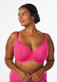 Rozā mežģīņu krūšturis ar platiem regulējamiem plecu siksnām, dekoratīvu apdari un ziedu rakstu uz kausiņiem. Apakšā gluda auduma apdare.