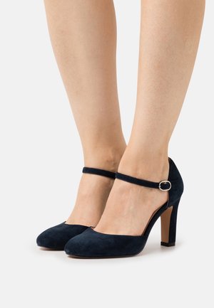Des pieds chaussés de chaussures à talons hauts Mary Jane en daim bleu marine avec des brides de cheville et une boucle en argent sur un fond blanc.