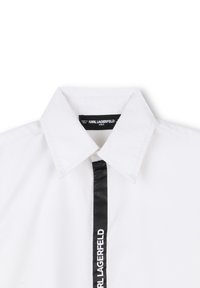 Hvit bomullsskjorte med strukturert krage og en svart satengstripe nedover foran som har "KARL LAGERFELD KIDS"-logoen.