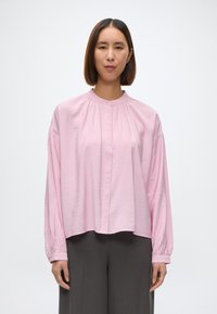 Lys rosa langermet bluse med rund hals, frontlukking med knapper og plisserte detaljer, kombinert med grå vide bukser.