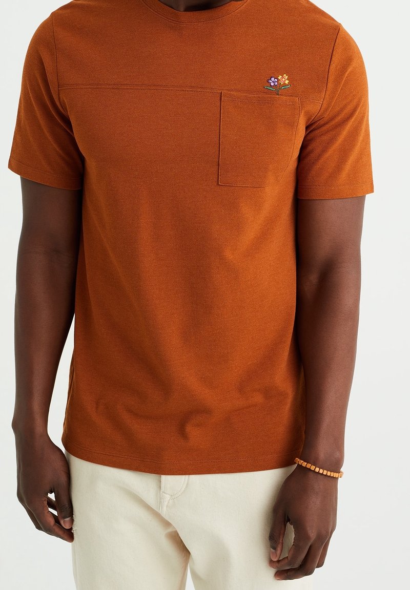 T-shirt a maniche corte, colletto rotondo, in tessuto arancione bruciato con una finitura testurizzata, dotato di una tasca applicata e ricami floreali.