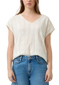Mujer que lleva una blusa blanca de encaje de manga corta con patrones florales metida en unos vaqueros azules, de pie con los brazos relajados.