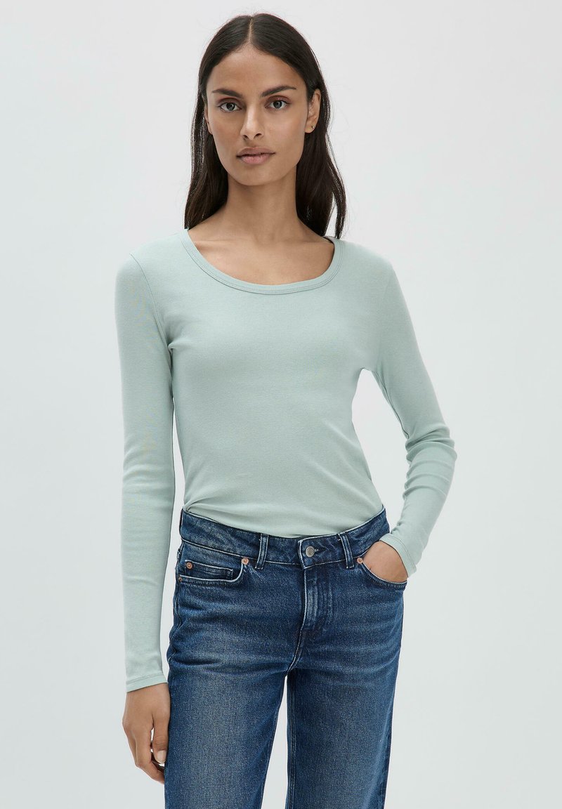 Maglietta a maniche lunghe di un verde menta chiaro, con scollatura a barca e una consistenza morbida e liscia. Abbinata a jeans di denim blu scuro.