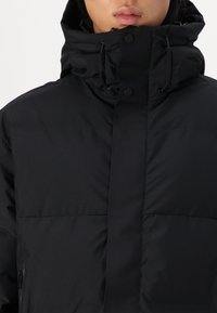 Nærbillede af en person iført en sort dunjakke med høj krave, trykknapper og justerbare snore til hætten, læber delvist synlige.