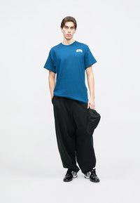 Μπλε βαμβακερό T-shirt με κοντά μανίκια, στρογγυλή λαιμόκοψη και λευκό λογότυπο. Μαύρες πλατιές παντελόνες, μαύρο καπέλο και μαύρα αθλητικά παπούτσια με ασημένιες λεπτομέρειες.