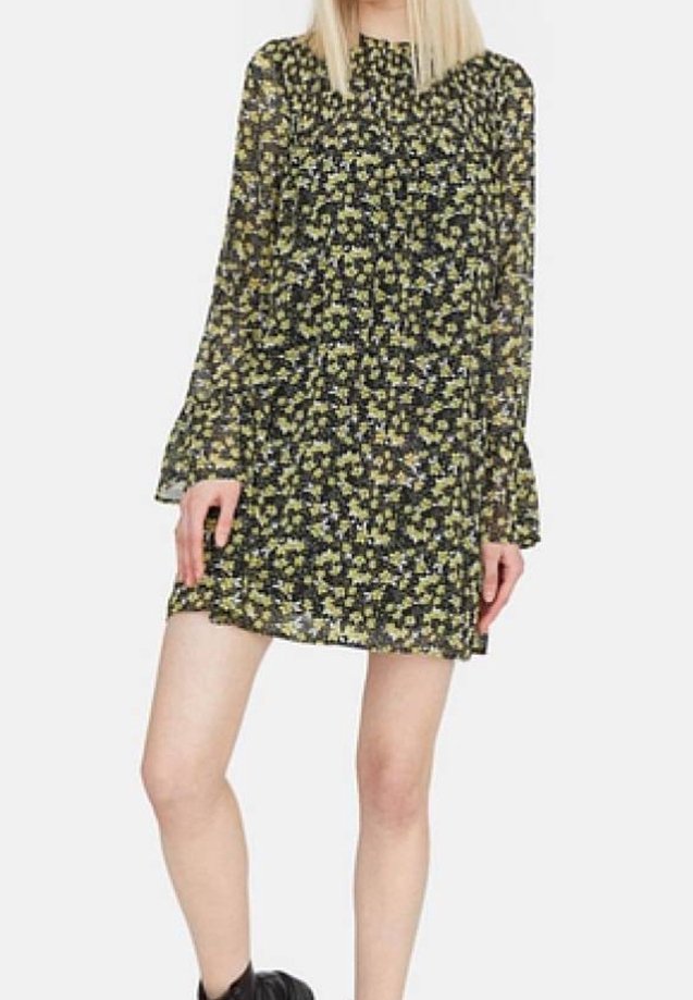 Femme vêtue d'une robe noire courte à manches longues avec un imprimé floral jaune et blanc et des bottines noires, debout les mains près des hanches.