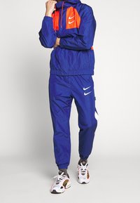 Námořnicky modrá větrovka s oranžovými doplňky, elastickými manžetami a kapucí; v kombinaci s odpovídajícími tepláky s bílým logem Nike a pruhy.