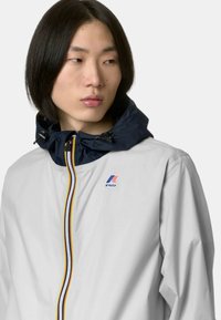 Giacca impermeabile grigio chiaro con cappuccio blu navy, zip gialla e blu a contrasto e logo K-Way sul petto. Texture liscia.