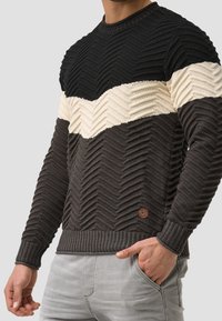 Strickpullover mit einem Fischgrätmuster in Schwarz, Creme und Dunkelgrau. Mit gerippten Bündchen und Saum sowie einem braunen Markenpatch am unteren Ende.