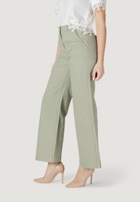 Pantaloni ampi di un verde chiaro in cotone con cuciture frontali sottili, abbinati a tacchi alti color nude e a un top bianco rifinito con pizzo.