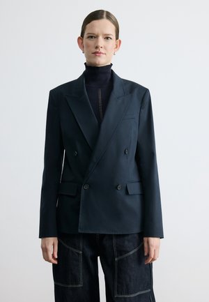 Naiste sassis juustega, kandes musta kahekordse rinnaosaga bleiserit musta kõrge kaelusega pluusi ja tumedate õmblustega pükstega, seisab valge tausta ees.