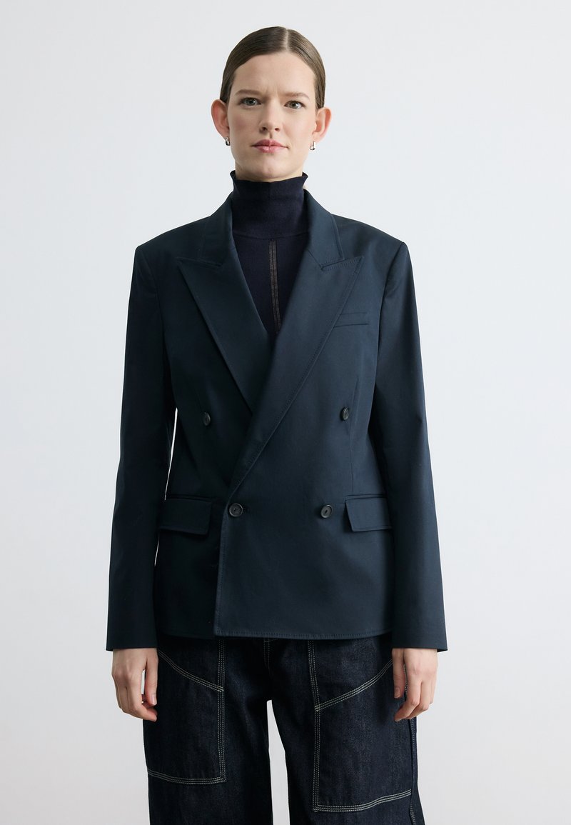 Femme aux cheveux lissés en arrière, portant un blazer noir croisé sur un col roulé noir et un pantalon sombre avec coutures visibles, debout devant un fond blanc.