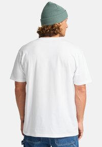 Hvid bomulds T-shirt med en afslappet pasform, korte ærmer og en glat tekstur. En lyseblå strikket hue bæres over krøllet hår.