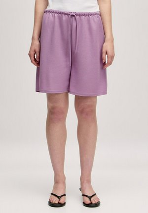 Lila sweatshorts met een elastische tailleband en verstelbaar trekkoord. Zachte textuur, knielengte en zijzakken. Gecombineerd met zwarte slippers.