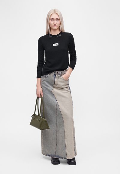 Jeune femme aux longs cheveux blonds portant un haut noir à manches longues, une longue jupe en denim bicolore, des chaussures noires et tenant un sac à main vert olive.