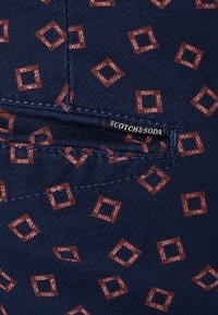 Navy blauwe stof met een geometrisch patroon van bruine vierkanten. Inclusief een klein label met "SCOTCH & SODA" in het wit. Gestructureerd oppervlak.